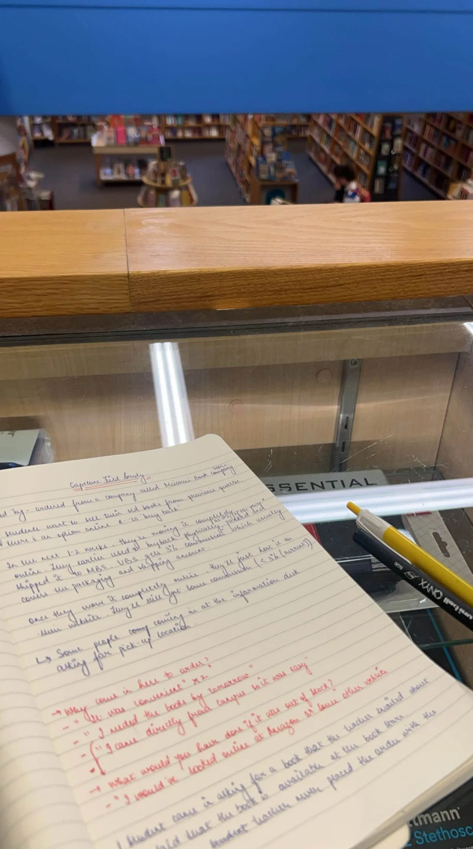 Field photograph from contextual inquiry: bookstore shelves seen over a counter, with a notebook of handwritten field notes and pens on the glass case below