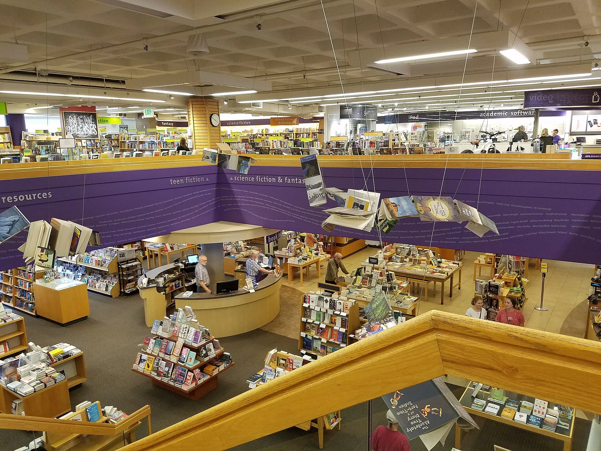 The University Book Store's main floor seen from an upper level: book tables, a round service desk with staff, and purple section signs
